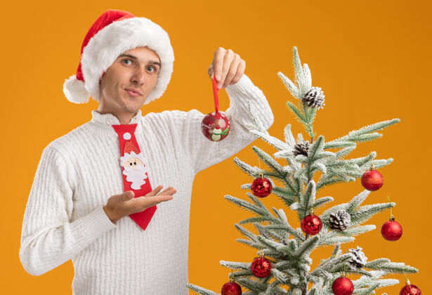 圣诞自信的年轻帅哥戴着圣诞帽 打着圣诞老人的领带 站在装饰好的圣诞树旁 手拿着圣诞球 看着橙色的墙上孤零零的装饰物指着小伙子圣诞老人