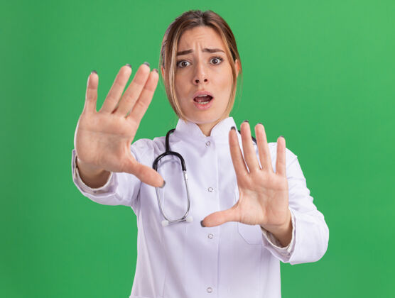 抱着年轻的女医生穿着医用长袍 手持听诊器 面对隔离在绿墙上的摄像机 吓得两手空空脸公民医学