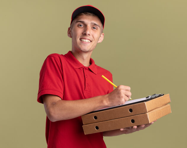 绿色微笑的年轻金发送货员拿着铅笔和剪贴板在比萨饼盒上持有金发微笑