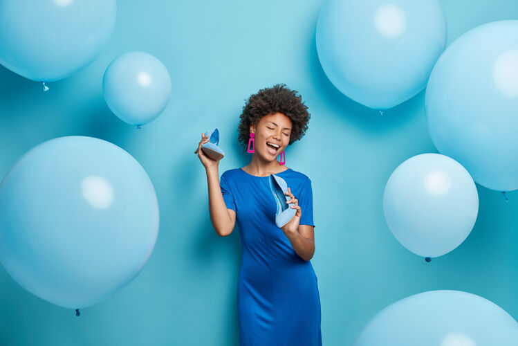 聚会卷发的女人把鞋子放在嘴边假装唱歌穿裙子为聚会选衣服蓝色隔离女人室内生日