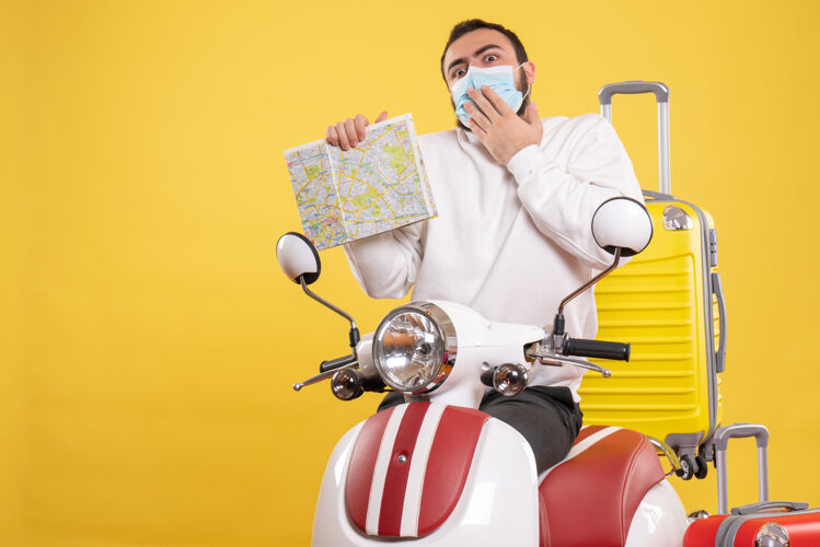 手持旅行概念的俯视图 戴着医用面罩的困惑的家伙站在摩托车旁边 黄色的手提箱在上面 手里拿着地图视图地图人
