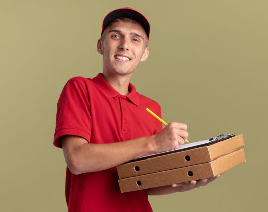 绿色微笑的年轻金发送货员拿着铅笔和剪贴板在比萨饼盒上持有金发微笑