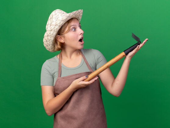 举行兴奋的年轻斯拉夫女园丁戴着园艺帽拿着耙子看着穿花园年轻