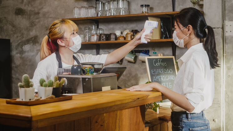 员工年轻的女餐厅工作人员戴着防护面罩 在进入前用红外线测温仪或测温枪对着顾客的额头出纳健康商务