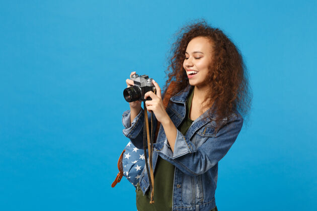 摄影师年轻的非洲裔美国女孩十几岁的学生在牛仔服装背包举行相机隔离在蓝色的墙上女性摄影女孩