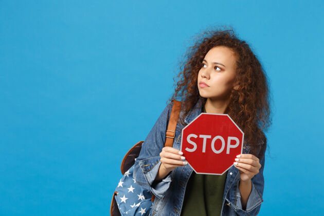 手势年轻的非洲裔美国女孩十几岁的学生在牛仔服装 背包举行停止孤立的蓝色墙壁站立人信息
