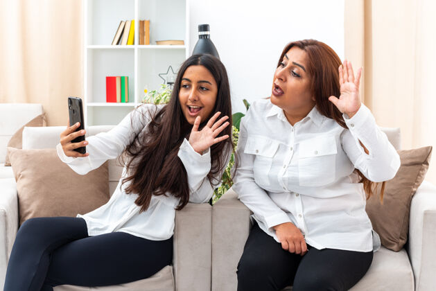 生活中年妇女和她的小女儿穿着白衬衫和黑裤子坐在椅子上用智能手机视频通话快乐和积极挥手在明亮的客厅轻椅子挥手