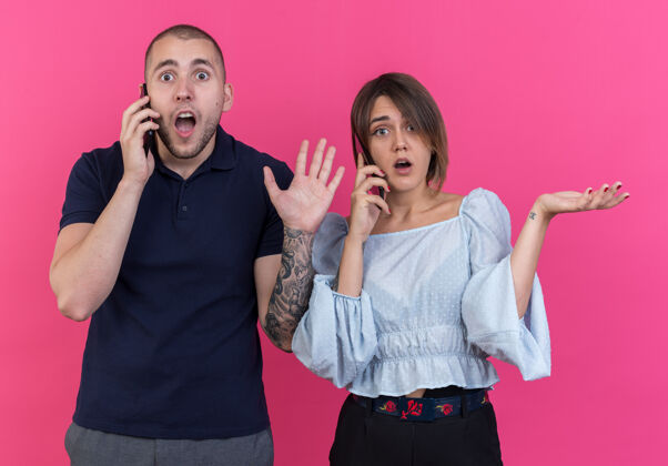 摆姿势年轻漂亮的夫妇站在粉红色的墙上 一边说着手机 一边举手 一边困惑着站着电话举起