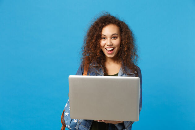 使用年轻的非洲裔美国女孩十几岁的学生在牛仔服装 背包工作的pc隔离在蓝色的墙壁上持有电脑青少年