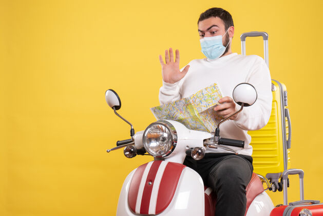 地图旅行概念的俯视图 戴着医用面罩的困惑的家伙坐在摩托车上 黄色手提箱在上面 手里拿着地图举行顶摩托车