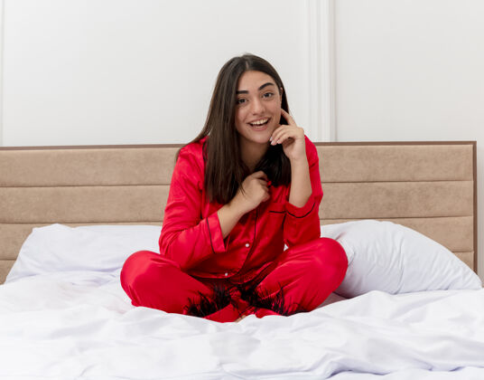 光穿着红色睡衣的年轻漂亮女人坐在床上放松地微笑着 在卧室里开心地笑着睡衣漂亮脸