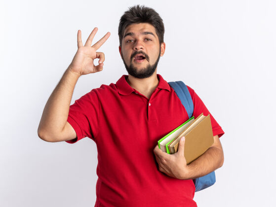 看年轻的留着胡子的学生 穿着红色马球衫 背着背包 手里拿着笔记本 看上去很开心 积极地在白墙上显示ok标志站立年轻姿势