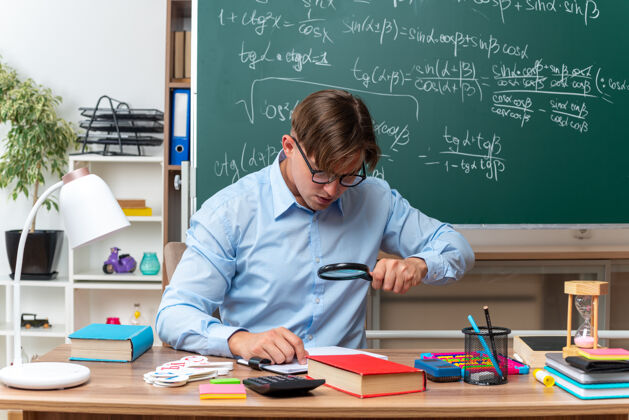 穿过年轻的男老师戴着眼镜通过放大镜看笔记备课坐在课桌旁 教室的黑板前放着书和笔记书桌教室笔记
