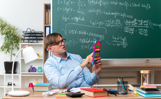 课年轻的男老师戴着眼镜 带着账单 在教室的黑板前拿着书和笔记坐在课桌旁备课账单笔记教室