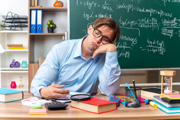 年轻戴着眼镜的年轻男老师又累又无聊 想坐在课桌旁睡觉 教室里黑板前放着书和笔记黑板睡眠教室