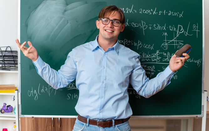 教室年轻的男老师戴着眼镜拿着海绵微笑着自信地站在教室里数学公式的黑板旁数学年轻人公式