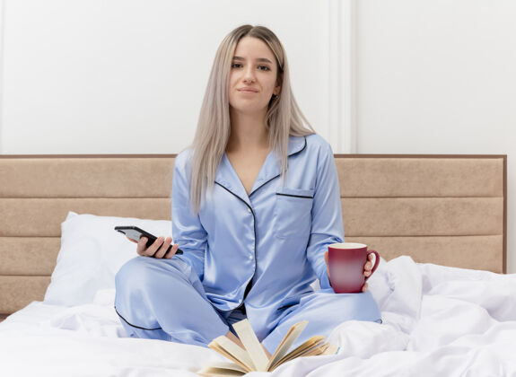 室内穿着蓝色睡衣的年轻美女坐在床上 用智能手机喝着咖啡 在卧室里微笑背景灯光使用