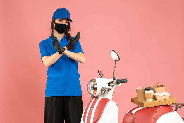 摩托车俯视图：戴着医用面罩的快递员女孩站在摩托车旁边 摩托车上放着咖啡蛋糕 在柔和的桃色背景下做出停车手势医疗背景男性