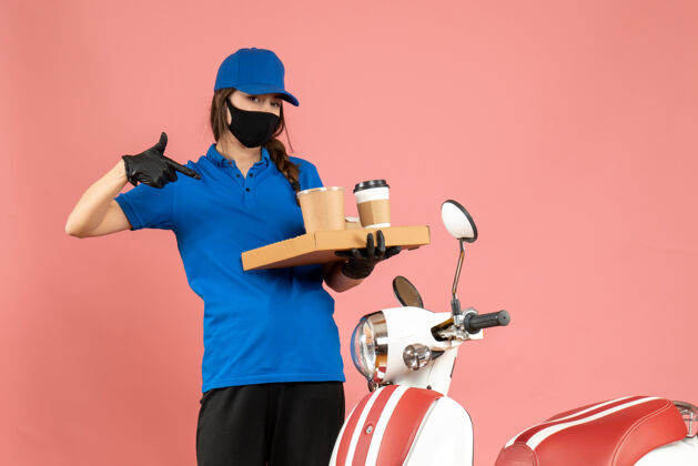 指俯视图：戴着医用口罩手套的快递员女孩站在摩托车旁指着粉色背景上的咖啡小蛋糕小面具专业