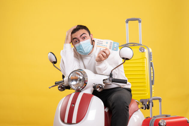 坐着旅行概念的俯视图 戴着医用面罩的年轻人坐在摩托车上 带着黄色手提箱 拿着车票感到困惑摩托车人抱着