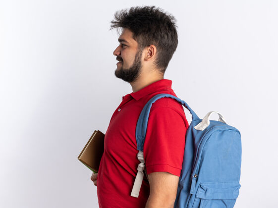 背包快乐的留着胡子的年轻学生 穿着红色马球衫 背着背包 拿着笔记本 横着站在白墙上人姿势马球