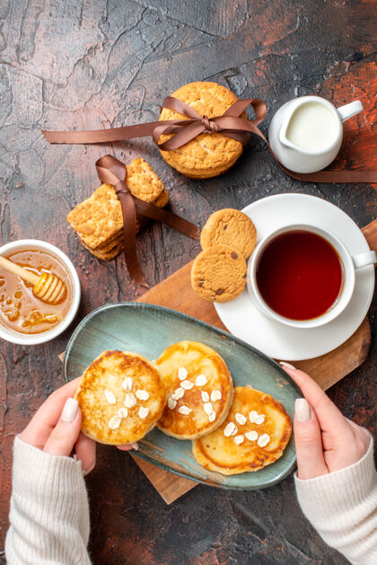 蜂蜜上图：手拿托盘和新鲜的薄煎饼一杯红茶放在木制砧板上蜂蜜饼干牛奶堆在黑暗的表面上晚餐盘子木板