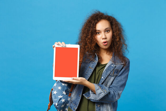 显示器年轻的非洲裔美国女孩十几岁的学生在牛仔服装 背包举行垫电脑隔离在蓝色的墙壁上设备空白促销