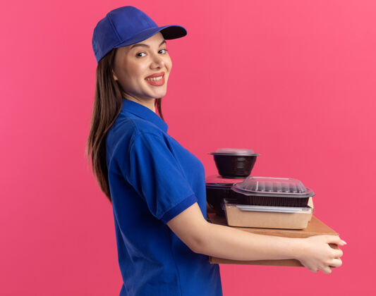 漂亮微笑着的漂亮送货员身着制服 站在一边 手里拿着比萨饼盒上的食品包和容器送货容器女人