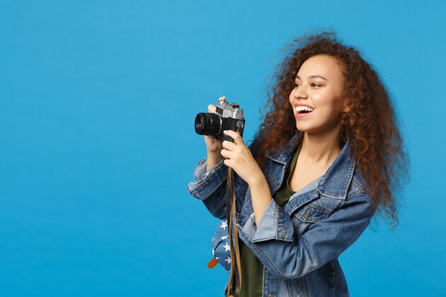 图片年轻的非洲裔美国女孩十几岁的学生在牛仔服装背包举行相机隔离在蓝色的墙上学生人民族