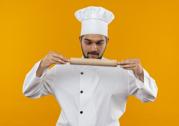 年轻印象深刻的年轻男厨师穿着厨师制服拿着擀面杖看着隔离在橙色墙上烹饪制服卷