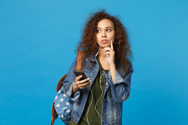 手机年轻的非洲裔美国少女 穿着牛仔服 背着耳机 隔离在蓝色的墙上举行女人