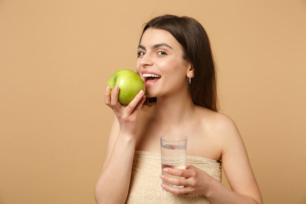 健康近距离拍摄完美肌肤的半裸女人 裸妆将苹果和水隔离在米色粉彩墙上身体护理纯净肩膀