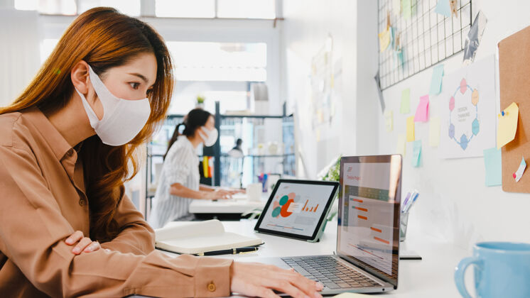 女性亚洲女企业家戴着医用面罩在新的正常情况下保持社会距离以防病毒 同时在办公室使用笔记本电脑背部亚洲人团队合作