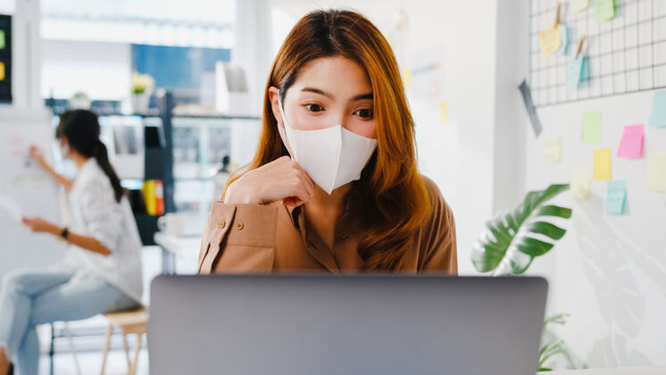 工作亚洲女商人戴口罩 在新的正常情况下保持社交距离 以防病毒 在办公室使用笔记本电脑向同事演示视频通话中的计划在线学生互联网