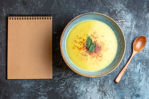 午餐俯瞰图螺旋笔记本美味的汤与胡椒和薄荷在一个蓝色的锅旁边的木勺在黑暗的背景黑碗胡椒