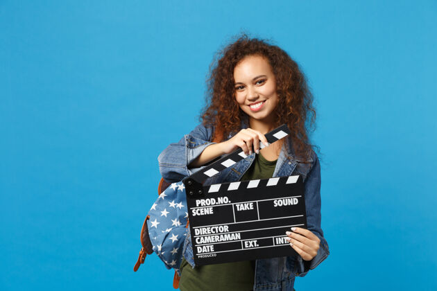 学生年轻的非洲裔美国女孩十几岁的学生在牛仔服装 背包举行克拉珀隔离在蓝色的墙壁上人黑板摄影