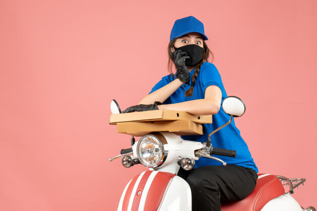 桃俯视图：戴着医用面罩和手套的年轻女快递员坐在滑板车上 用粉彩桃子送订单运动员射击粉彩