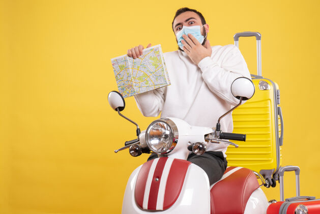 手持旅行概念的俯视图 戴着医用面罩的困惑的家伙站在摩托车旁边 黄色的手提箱在上面 手里拿着地图视图地图人