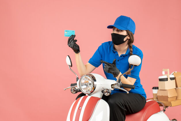 粉彩顶视图自信的女快递员戴着医用面罩和手套坐在踏板车上拿着银行卡 在柔和的桃色背景下传递订单穿背景桃
