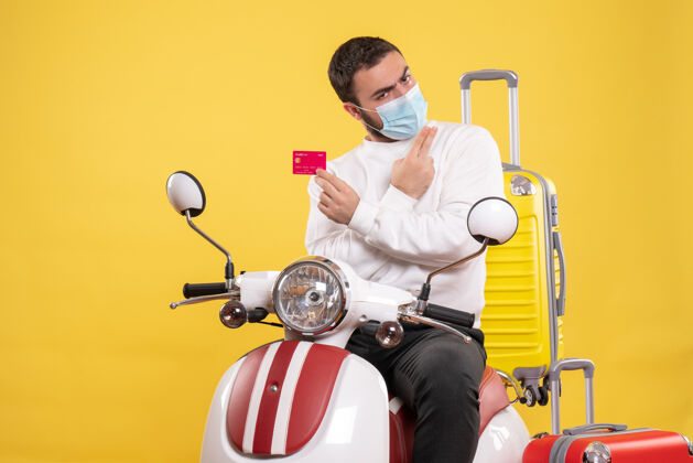 前面旅行概念的正面图 戴着医用面罩的自信年轻人坐在摩托车上 黄色手提箱上 手里拿着银行卡银行医疗摩托车