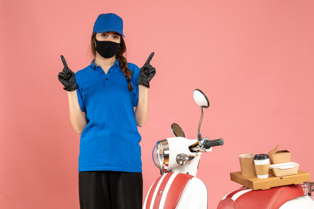 背景顶视图自信的信使女孩戴着医用口罩手套站在摩托车旁边 咖啡蛋糕在粉彩桃色背景上指向两边摩托车蛋糕穿