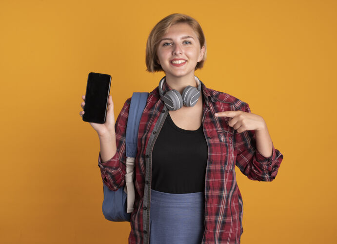 点微笑的年轻斯拉夫女学生戴着耳机背着背包 指着电话空间耳机电话