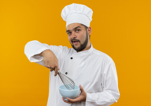 厨师令人印象深刻的年轻男性厨师在厨师制服拿着威士忌和碗隔离在橙色墙上持有男性制服