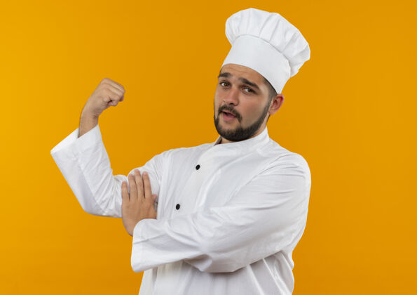 手势自信的年轻男厨师身着厨师制服 动作有力 抚摸着隔离在橙色墙上的肌肉男性触觉自信