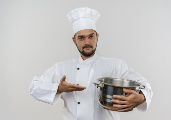 自信身着厨师制服的自信年轻男厨师手拿并指着隔离在白墙上的锅年轻烹饪厨师