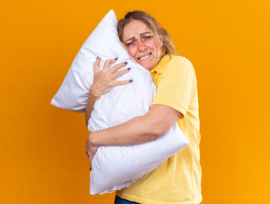 不健康穿着黄色衬衫的不健康的女人感觉身体不适患流感 站在橘黄色的墙上抱着发烧的抱枕感觉感冒痛苦