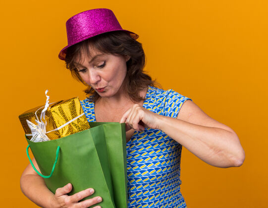 年龄中年妇女戴着派对帽 手里拿着装有生日礼物的纸袋 站在橘色的墙上兴致勃勃地庆祝生日派对生日举行帽子
