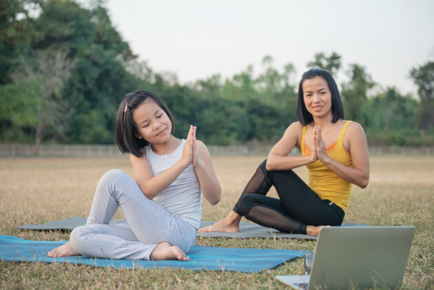 积极妈妈和女儿做瑜伽妇女和儿童在公园里训练户外运动健康的运动生活方式 看瑜伽练习在线视频教程和伸展在阿达松手姿势伸展孩子电脑