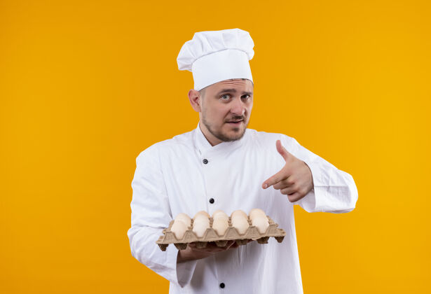 纸箱自信的年轻帅哥厨师穿着厨师制服 拿着并指着隔离在橙色墙上的一盒鸡蛋年轻帅气厨艺