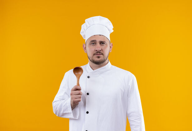手握自信的年轻帅哥厨师身着厨师制服 拿着木汤匙孤立在橙色的墙上勺子制服年轻
